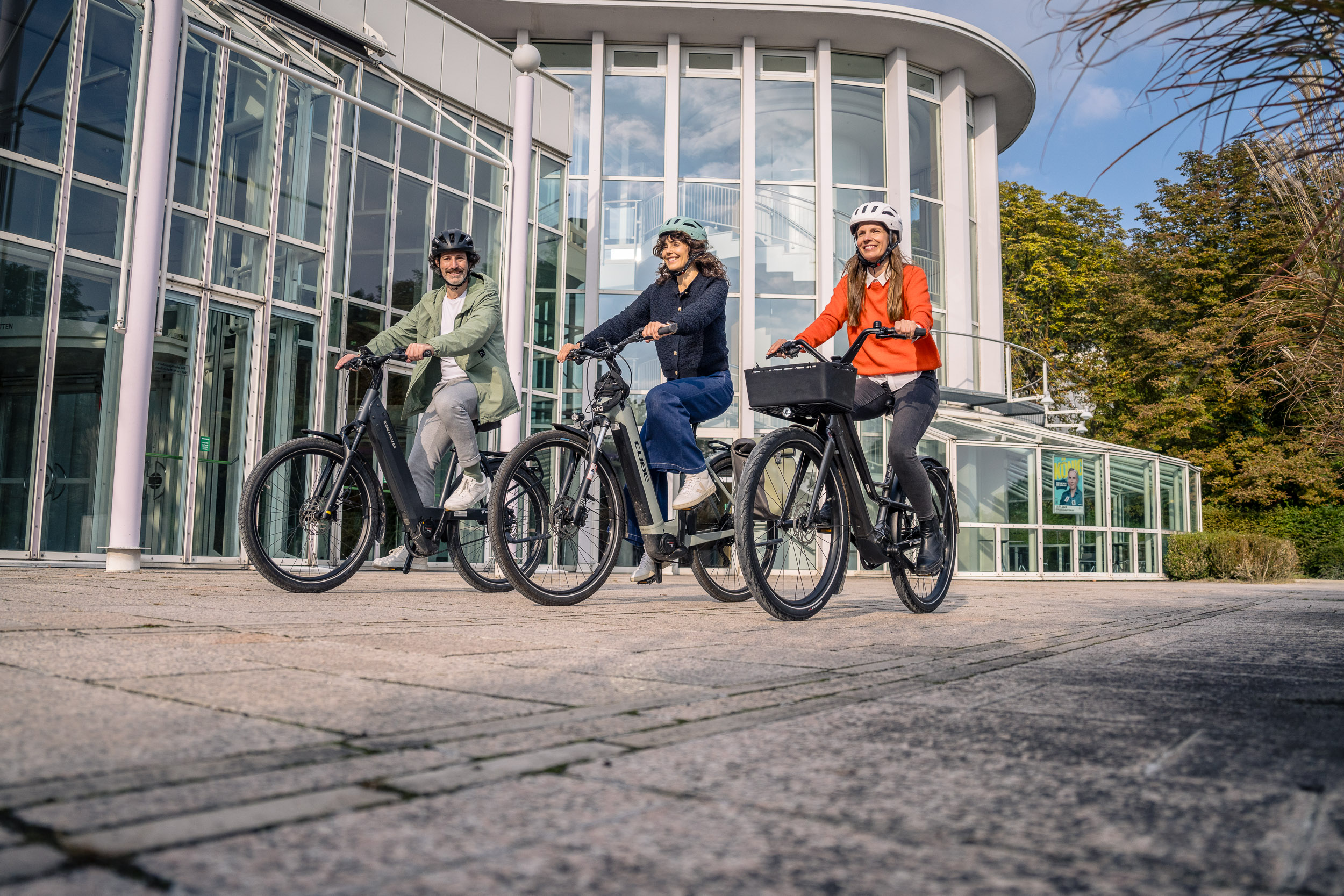 Drei Personen fahren mit Fahrrädern vor einem modernen Glasgebäude über einen gepflasterten Platz.