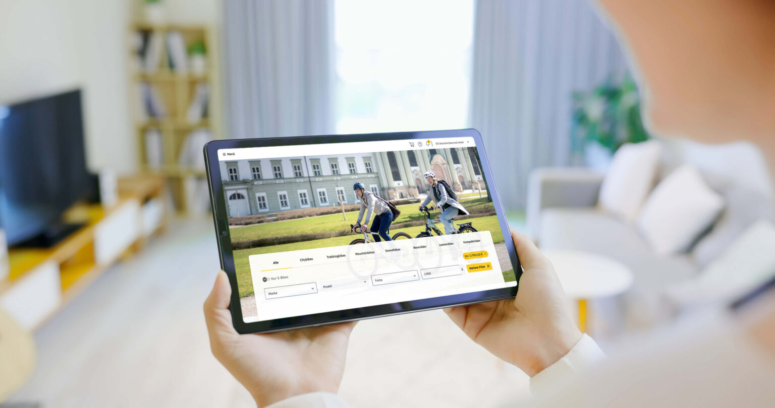 Eine Person hält ein Tablet, auf dem eine Webseite mit Fahrradauswahl und zwei Radfahrern im Freien angezeigt wird. Im Hintergrund ist ein modernes Wohnzimmer zu sehen.