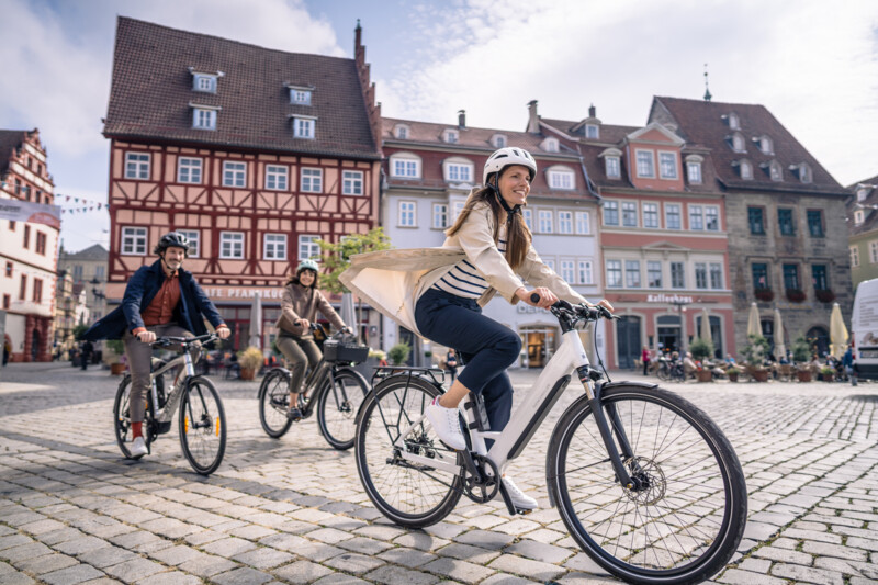 Drei Personen fahren mit Fahrrädern über einen Kopfsteinpflasterplatz vor historischen Fachwerkhäusern.