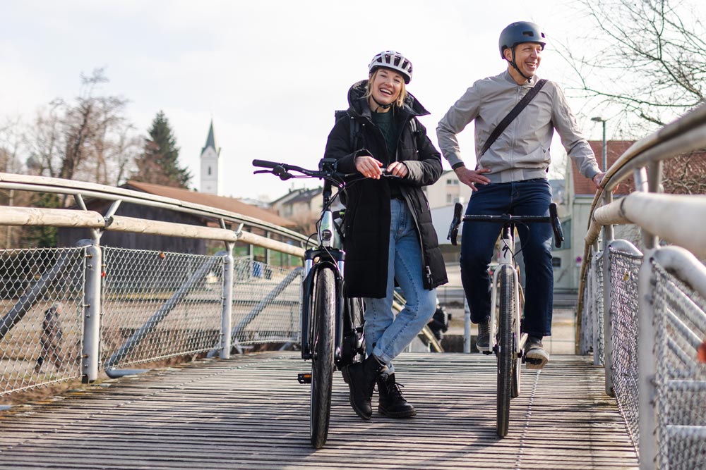 Ein Mann und eine Frau mit Helmen stehen lächelnd nebeneinander auf einer Brücke, während sie ihre Fahrräder halten. Im Hintergrund ist ein Kirchturm und eine ländliche Umgebung zu sehen.