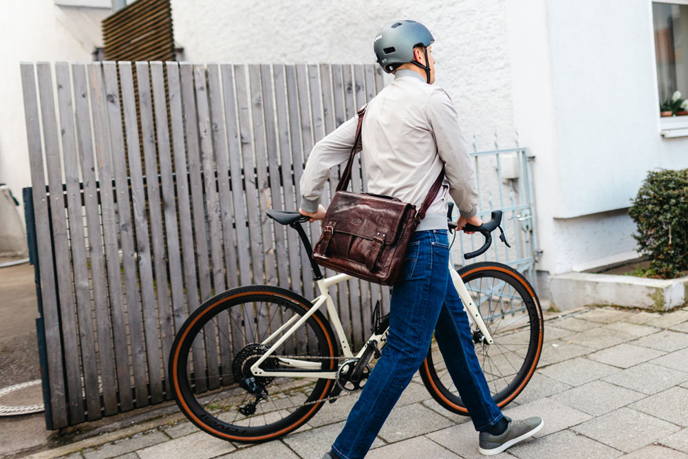 Ein Mann mit Fahrradhelm und Umhängetasche führt sein Rennrad einen Gehweg entlang, vorbei an einem Holzzaun.